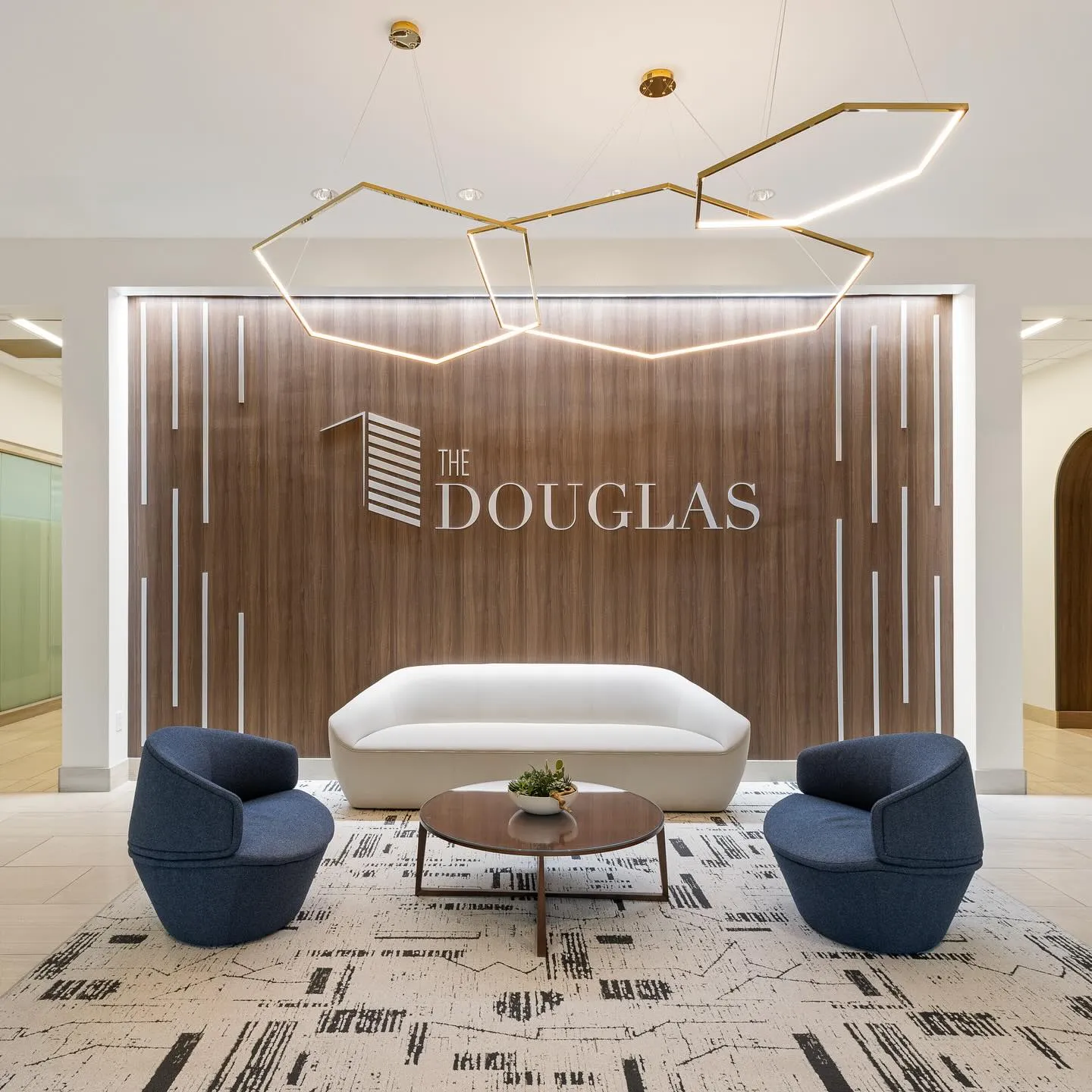 Modern lobby by Northgate Construction with a wood accent wall displaying The Douglas sign, white sofa, two blue chairs, a round coffee table, geometric light fixture, and a patterned rug on a tiled floor.
