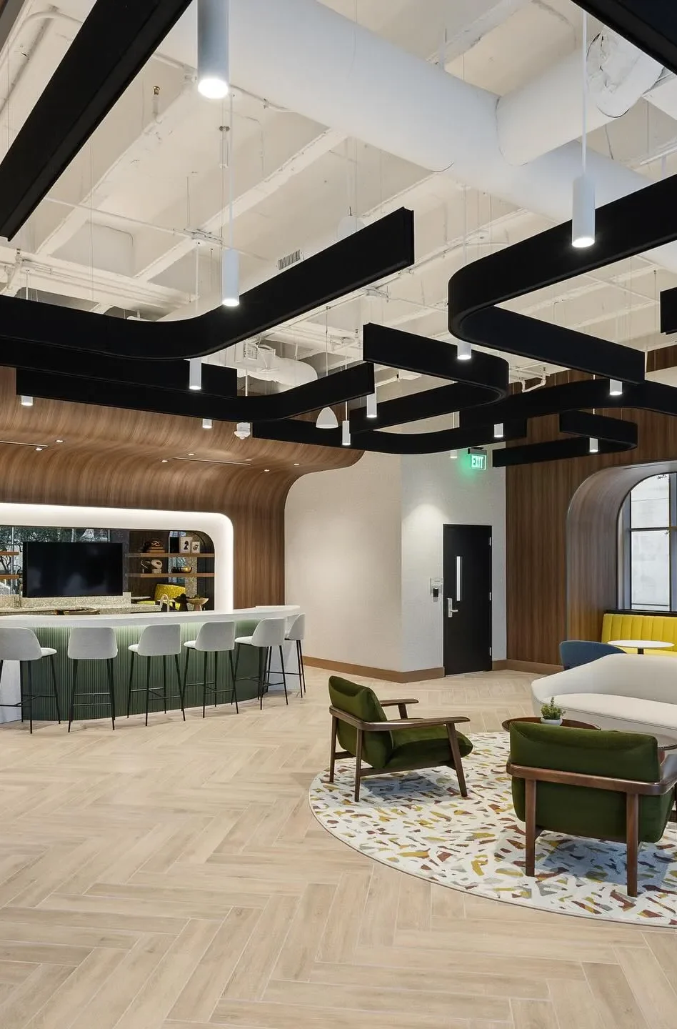 Modern office lounge by Northgate Construction featuring a curved wooden wall, bar seating at a counter, green armchairs around a round table on a patterned rug, and black ceiling beams with hanging lights.