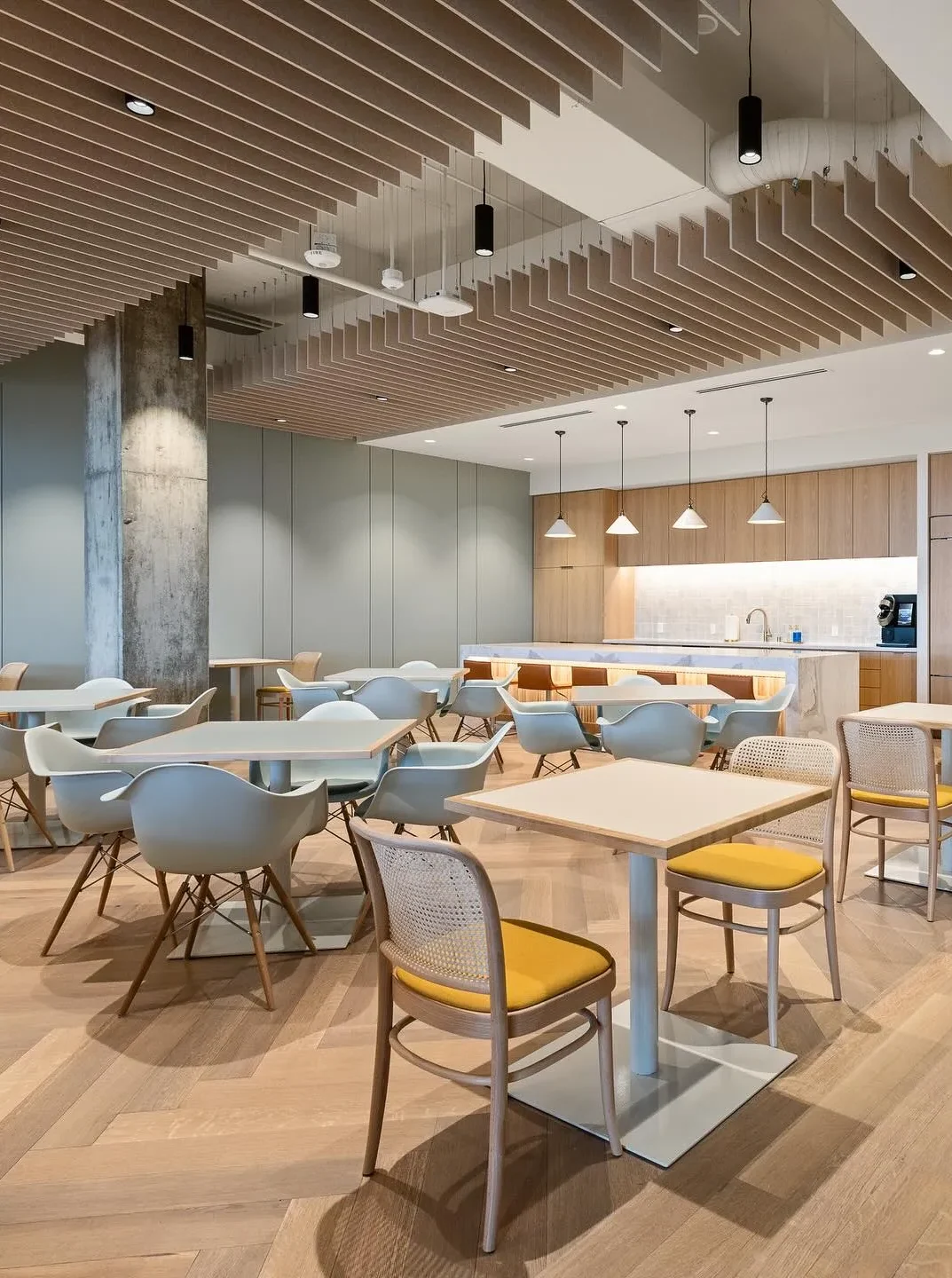 Modern office break room by Northgate Construction with light wood floors, contemporary tables and chairs, a kitchenette with pendant lights, and slatted wooden ceiling panels. The space is bright and neatly organized.