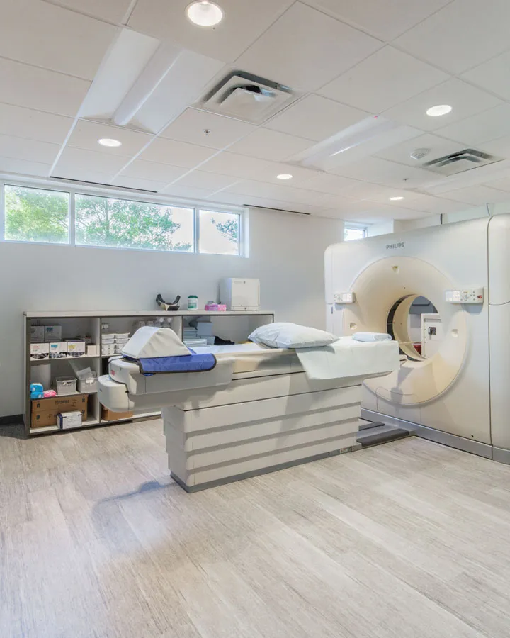 A modern medical imaging room, expertly crafted by Northgate Construction, features a large MRI or CT scanner, an examination table, shelves with medical supplies, and bright natural light from windows. The space is clean and organized.