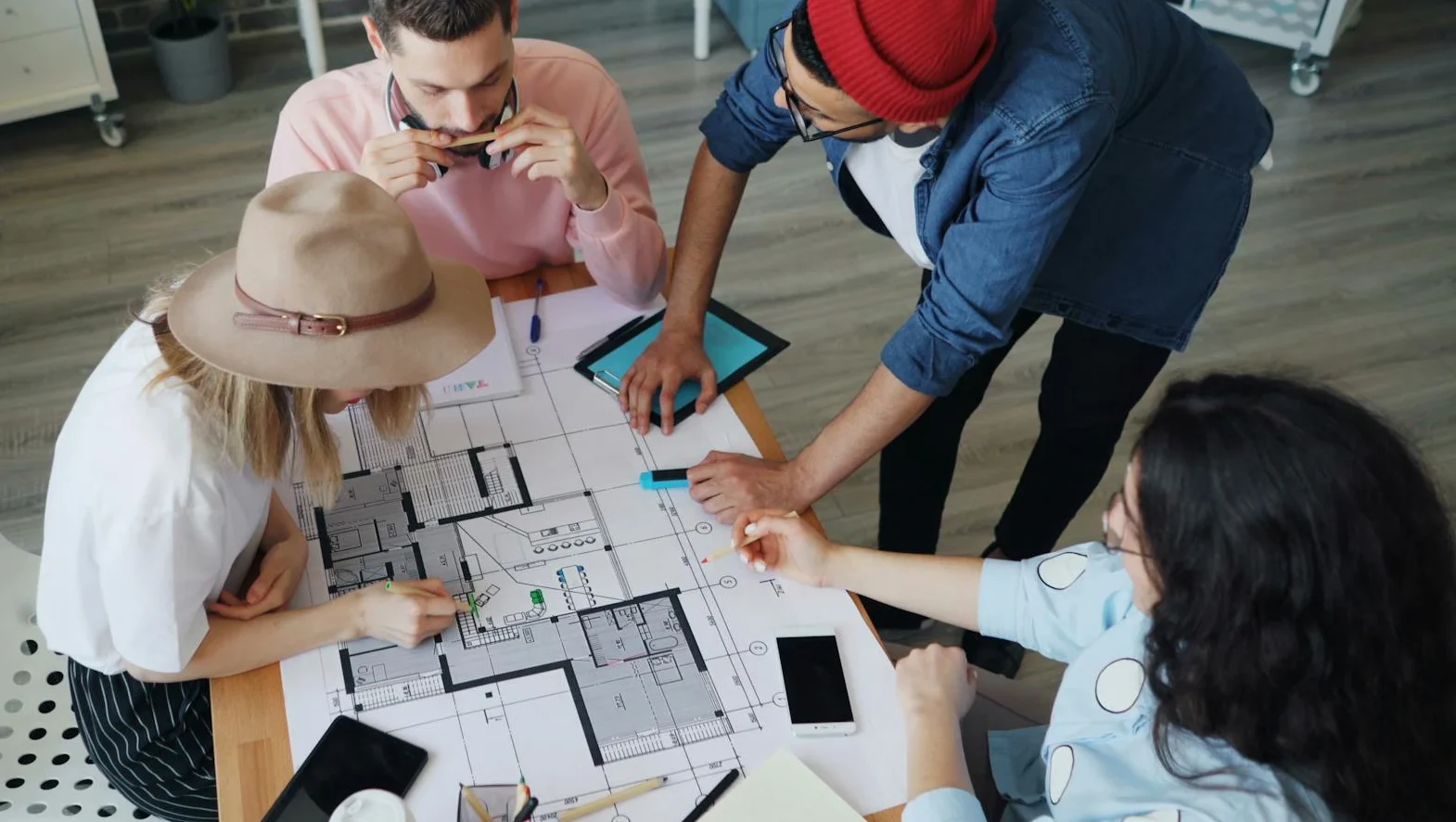 Four people gather around a table at Northgate Construction, examining architectural blueprints. Various devices, notebooks, and coffee cups are scattered around as one person points at the plans while others discuss ideas.