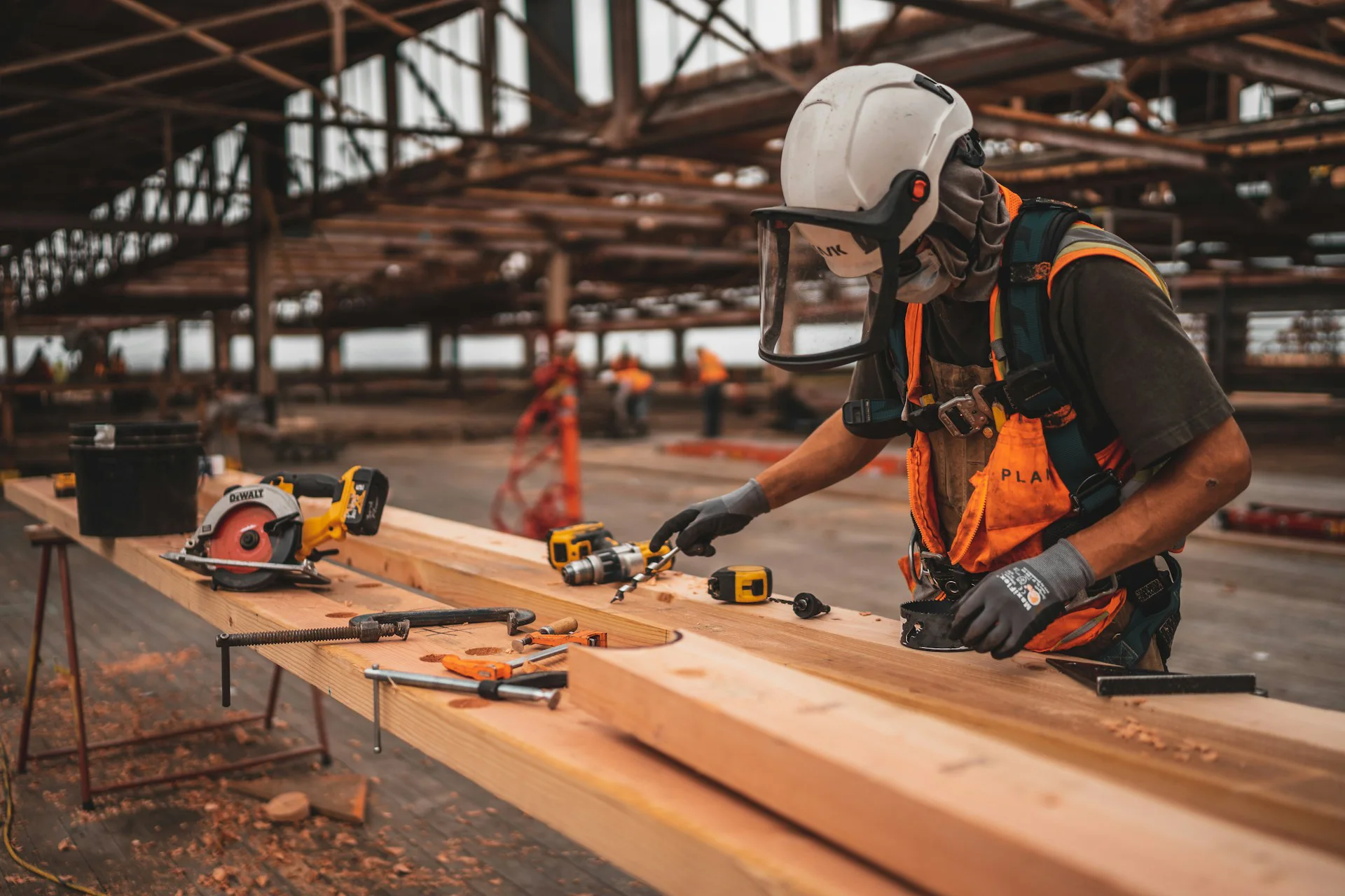 A Northgate Construction worker wearing safety gear uses power tools to work on wooden beams at a large indoor job site, with tools and equipment scattered on the workbench.