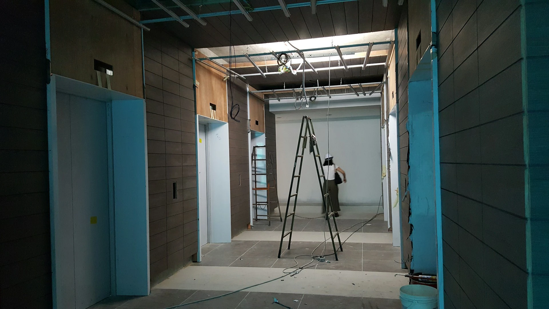 A person stands on a ladder under exposed ceiling wiring in a hallway under construction by Northgate Construction, flanked by elevator doors and brown wall panels, with construction materials scattered around.