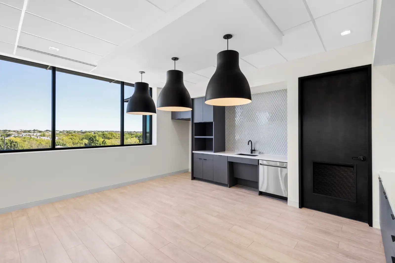 Modern office kitchenette by Northgate Construction with light wood floors, dark cabinets, a sink, dishwasher, three large black pendant lights, and a large window showcasing a scenic outdoor view.