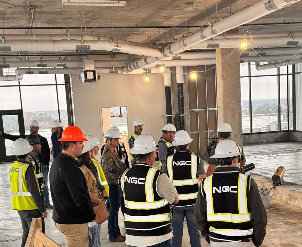 A group of northgate construction workers and supervisors wearing hard hats and safety vests stand inside a partially finished building with exposed ceilings and large windows.