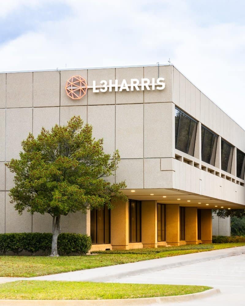 A modern beige office building, built by Northgate Construction, with large windows, the L3HARRIS logo and emblem on the corner, a tree and neatly trimmed bushes in front, and a partly cloudy sky above.