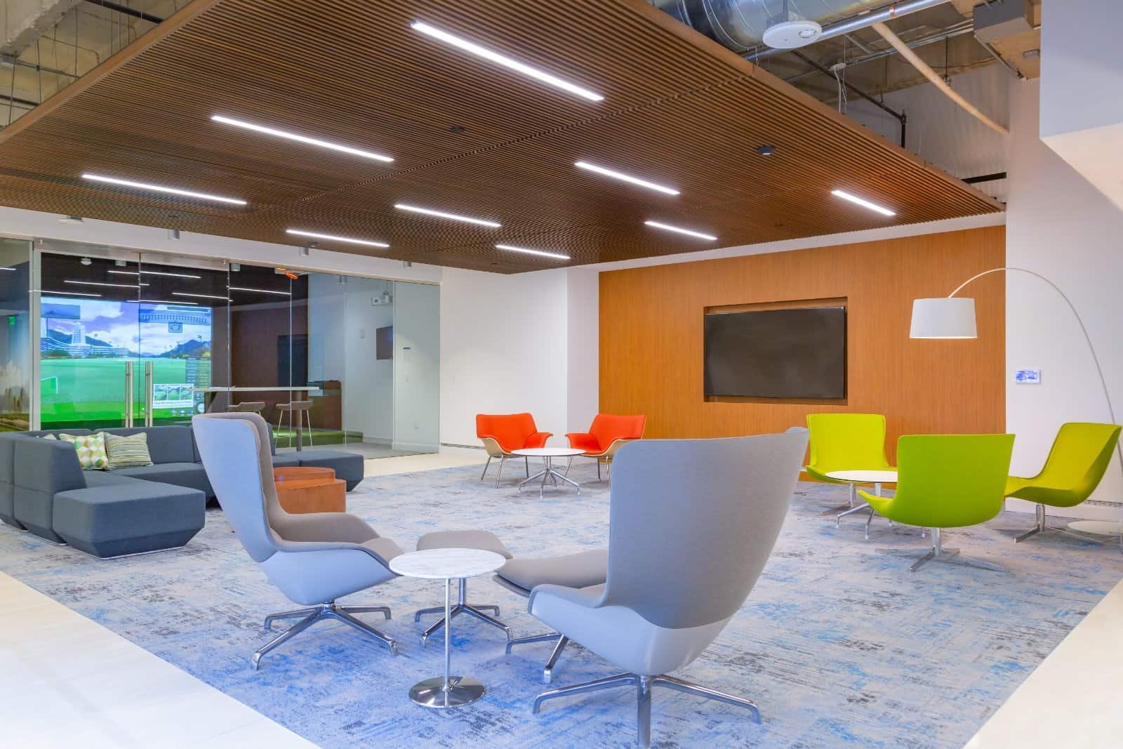 Modern office lounge by Northgate Construction with gray, green, and orange chairs, a blue patterned rug, small tables, and a wooden accent wall with a large screen. Ceiling has wooden slats and LED lights; glass wall shows a hallway.