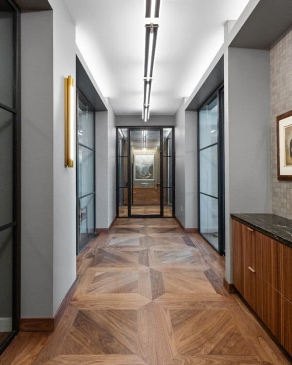 A modern hallway by Northgate Construction features geometric wooden flooring, gray walls, glass doors, a black countertop on the right, artwork on the walls, and ceiling lights running the length of the corridor.