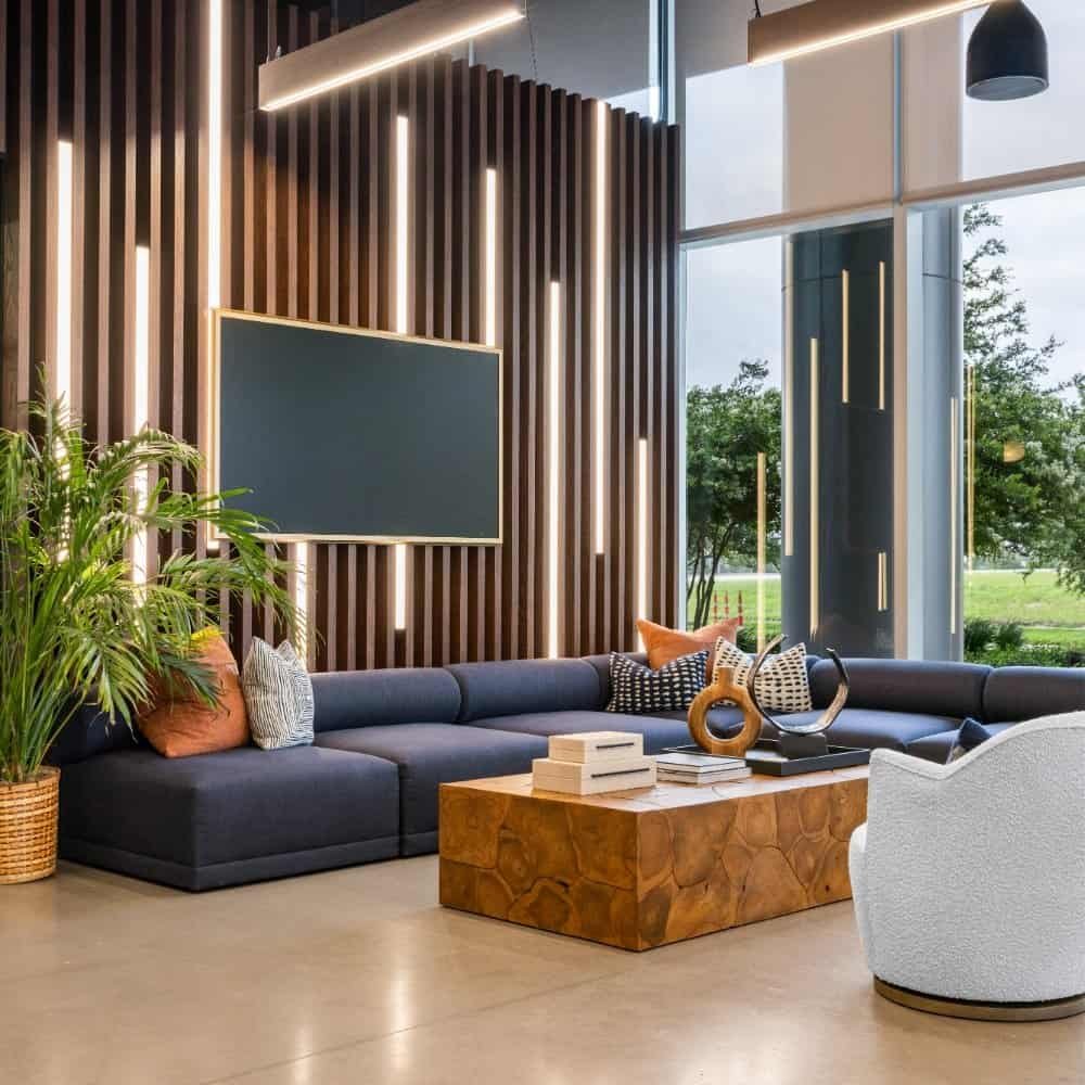 Modern lounge by Northgate Construction with a large navy sectional sofa, wood accent wall featuring vertical lights, a blank TV screen, geometric wooden coffee table, white swivel chair, and large windows revealing trees outside.