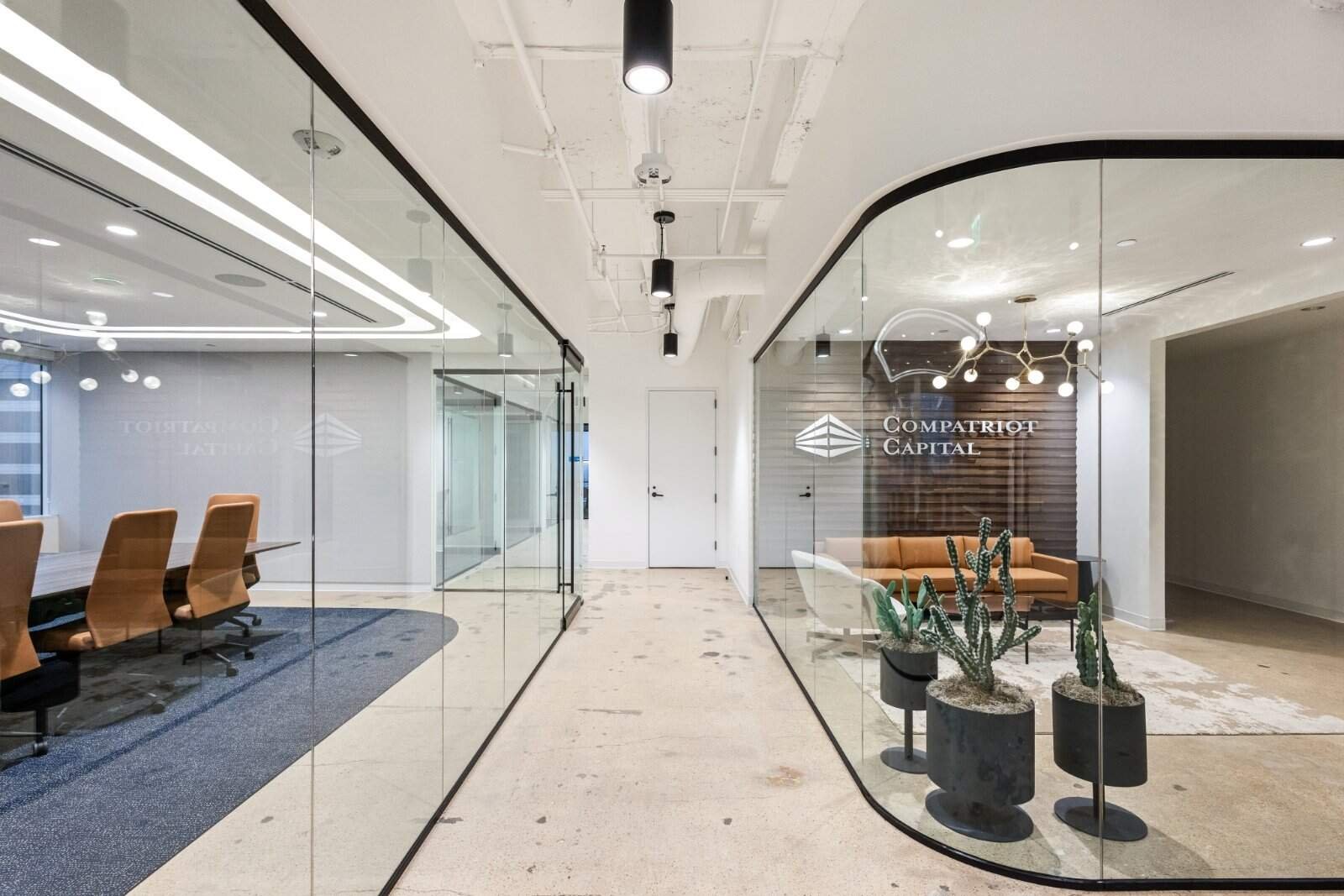 Modern office interior with glass walls, featuring a conference room with tan chairs on the left and a waiting area with a brown sofa and potted cacti on the right, thoughtfully crafted by Northgate Construction. Compatriot Capital sign is on the wall.