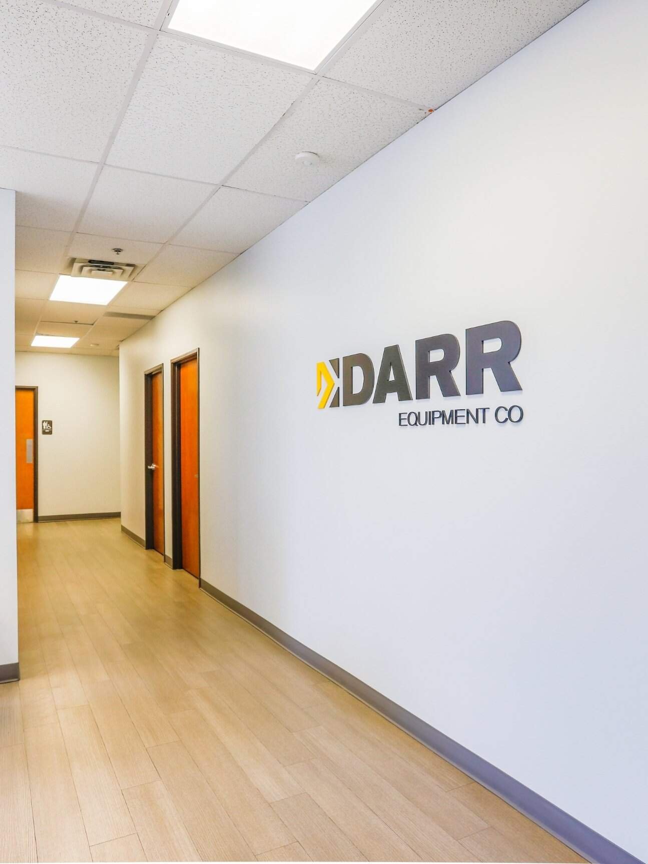 A clean, modern office hallway with light wood floors, white walls, and three closed wooden doors. DARR EQUIPMENT CO is displayed in large letters on the right wall, highlighting quality by Northgate Construction. Ceiling lights illuminate the space.