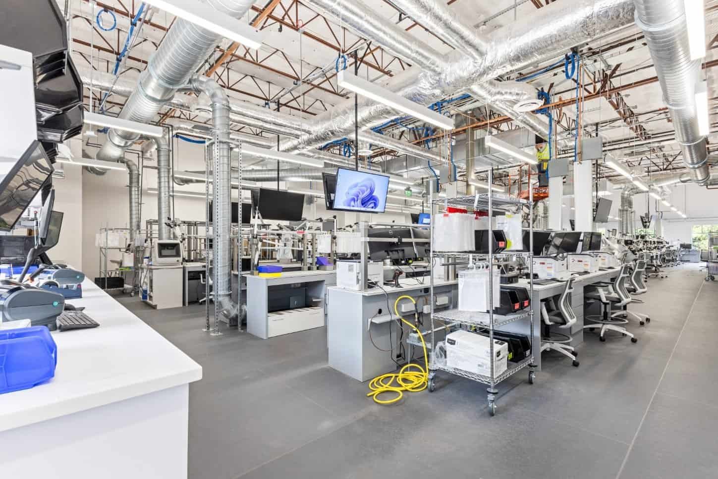 A spacious, modern laboratory built by Northgate Construction features white workstations, multiple computer monitors, and silver air ducts on the ceiling. Carts with equipment, chairs, and yellow cables fill the bright, organized space.