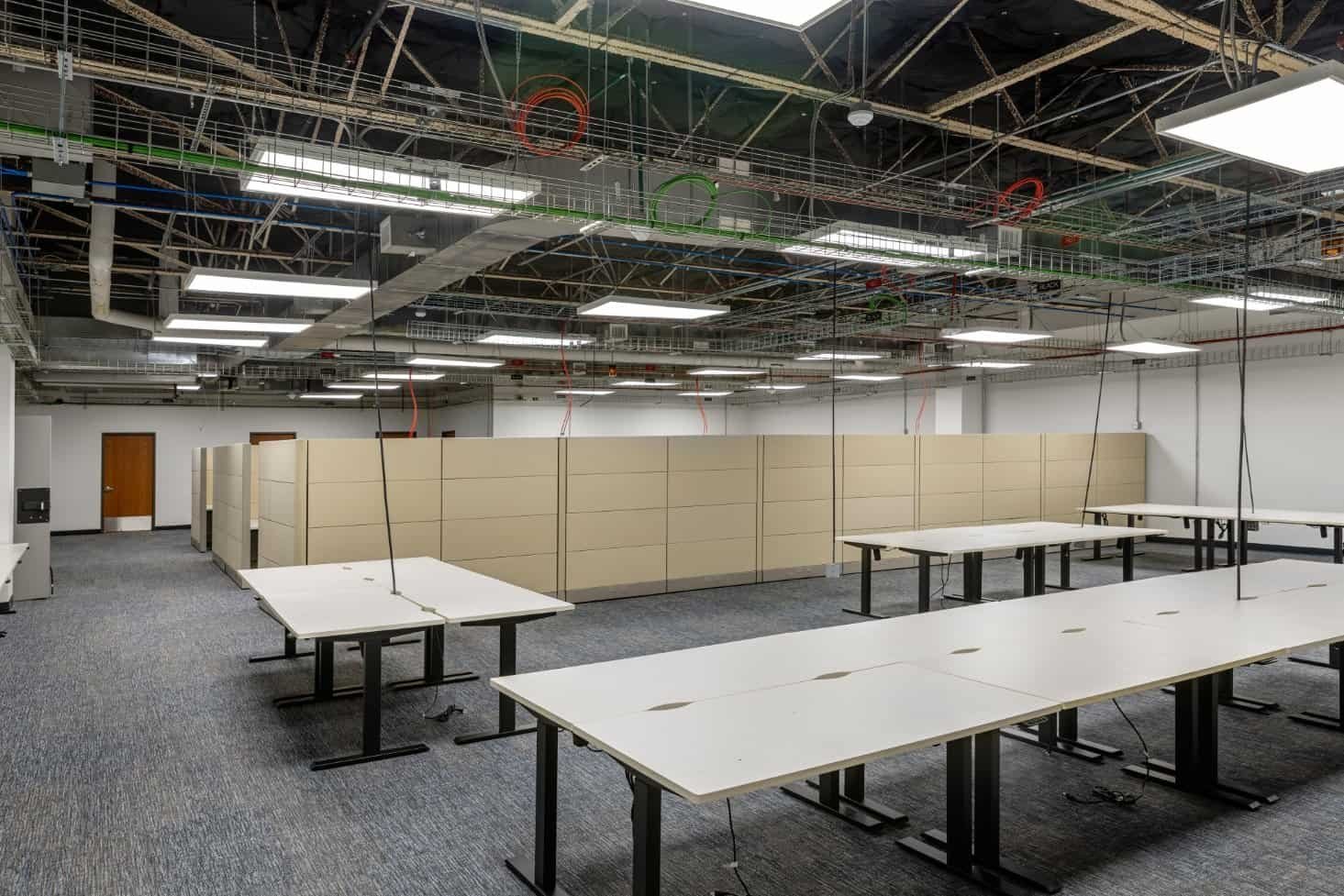 An office space under renovation by Northgate Construction, featuring exposed ceiling wiring, fluorescent lights, beige cubicles, and several white desks without chairs. The carpeted floor and plain white walls complete the scene.