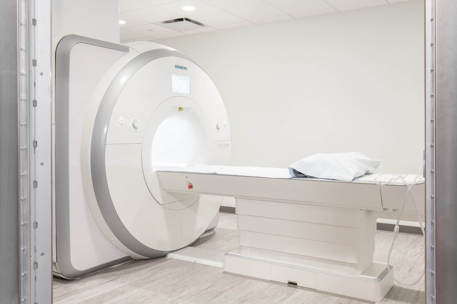 A modern MRI machine in a clean, bright medical room with a padded table and pillow, ready for a patient to undergo a scan. The machine is white with silver accents and has a digital display.