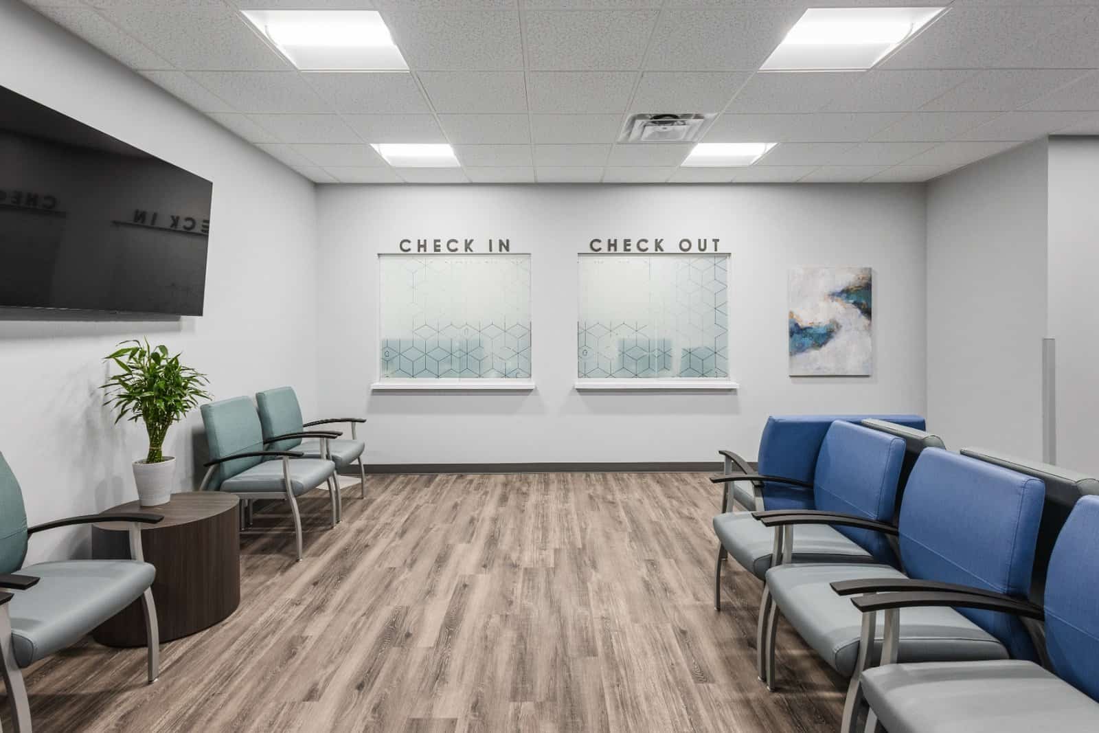 A clean, modern waiting room with blue chairs, wood flooring, a potted plant, a TV, and two labeled windows for check-in and check-out on the far wall. A piece of abstract art hangs on the right wall.