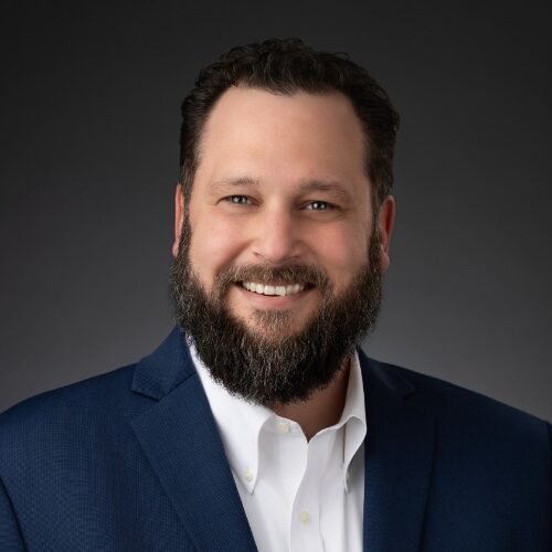 A man with a dark beard and short hair smiles at the camera. He is wearing a navy blue suit jacket over a white collared shirt, posing against a dark gray background.
