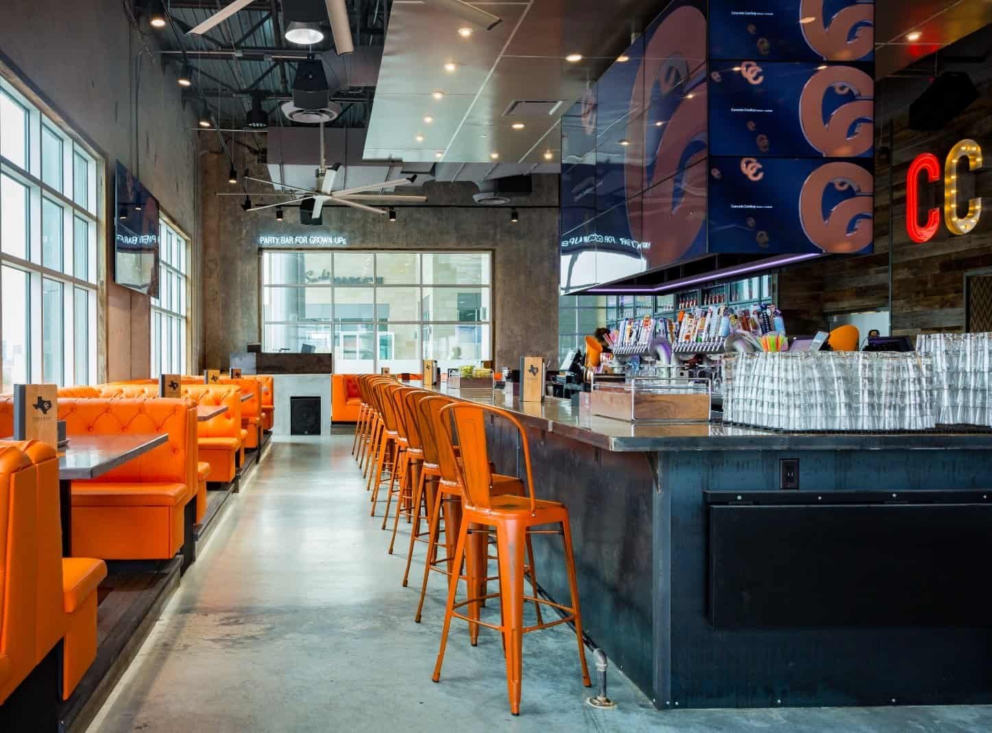 Modern restaurant interior with orange booths and matching bar stools, sleek concrete floors, a long bar with glasses stacked on it, and large windows allowing natural light to fill the space.