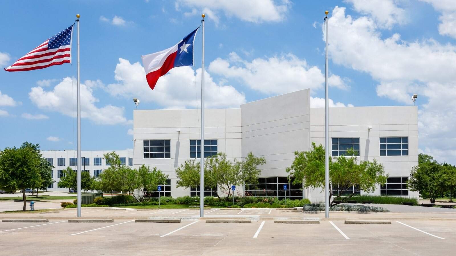 A modern white office building with American and Texas flags on flagpoles in front, surrounded by trees and an empty parking lot under a partly cloudy blue sky.