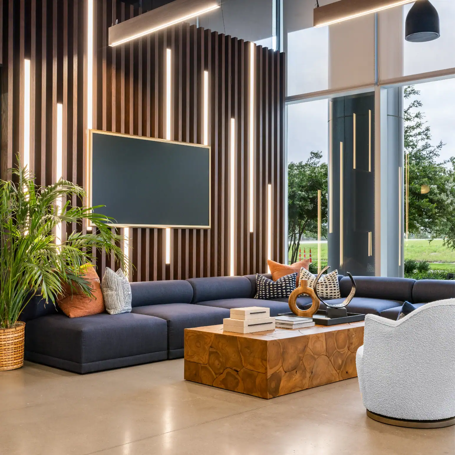 Modern lounge with a dark sectional sofa, decorative pillows, a wooden coffee table, and a potted plant. Vertical wood paneling with built-in lights and large windows with a view of greenery complete the stylish interior.