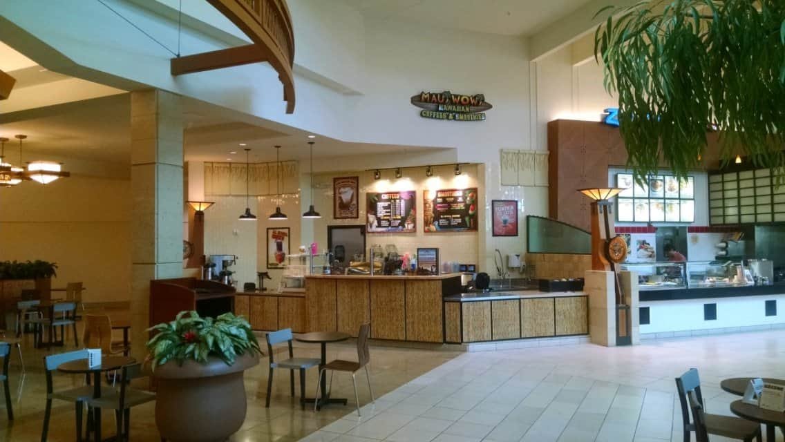 A nearly empty mall food court with several food stalls, including Maui Wowi and another with menu boards, surrounded by empty tables and chairs, potted plants, and bright natural light from windows.