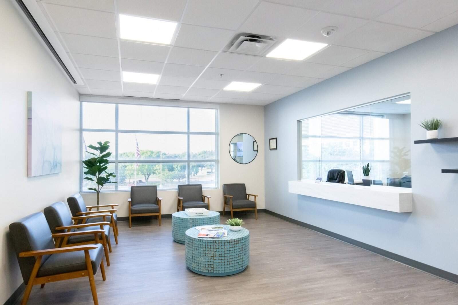 A modern waiting room with gray chairs, two round blue tables, potted plants, wall art, and a large window letting in natural light. A reception window is on the right wall.