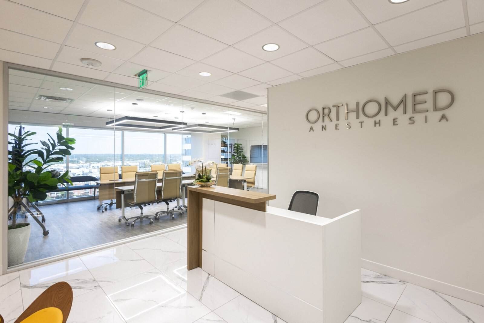 A modern office reception area with a desk, the sign OrthoMed Anesthesia on the wall, large white floor tiles, potted plants, and a glass-walled conference room with a long table and chairs in the background.