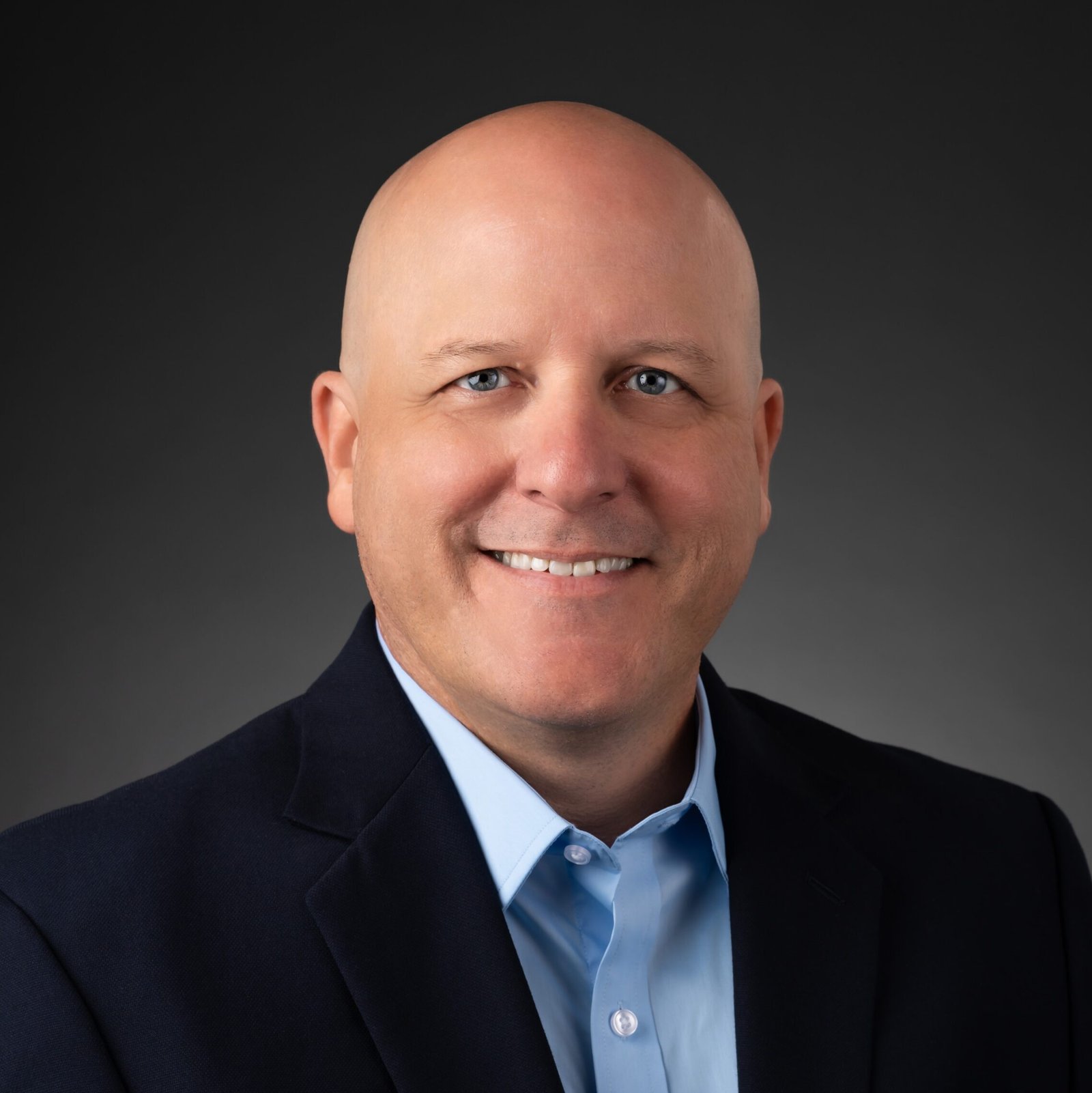A smiling bald man wearing a dark blazer and light blue dress shirt poses for a professional headshot against a dark, gradient background.