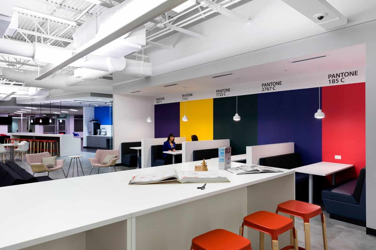 A modern office space with colorful Pantone-themed walls, white tables, and booths. Red stools line a counter, while a person sits working at a booth. Pendant lights hang from the ceiling, and shelves and seating are visible in the background.