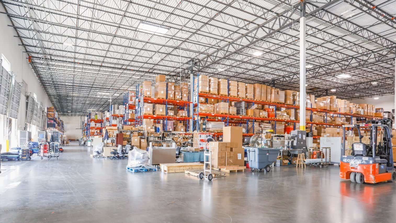 A spacious warehouse with high shelves stacked with boxes and goods, organized on orange racks. Forklifts and carts are scattered throughout the clean, well-lit space.