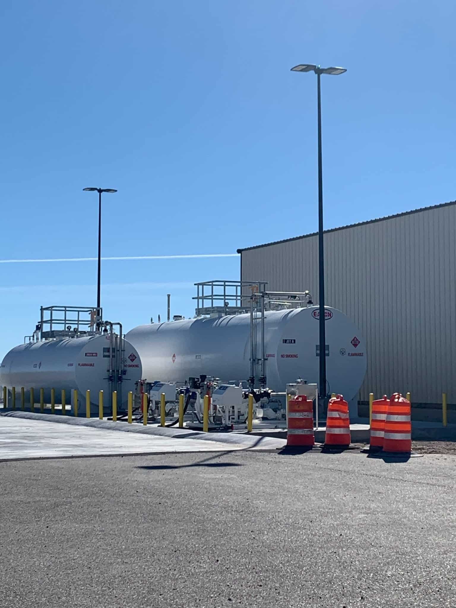 Two large white fuel storage tanks sit beside a beige industrial building under a clear blue sky. Orange traffic barrels and yellow safety bollards are positioned in front of the tanks.