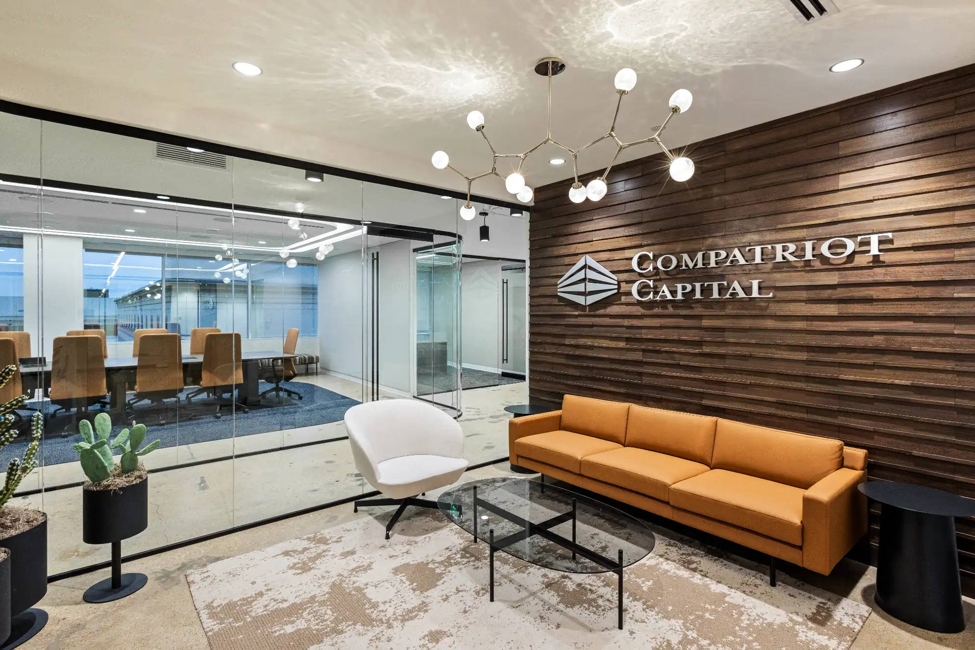 Modern office lobby with a yellow sofa, white chair, and glass coffee table. Wooden accent wall displays Compatriot Capital logo. Glass walls reveal a conference room with tan chairs and a large table. Contemporary lighting overhead.