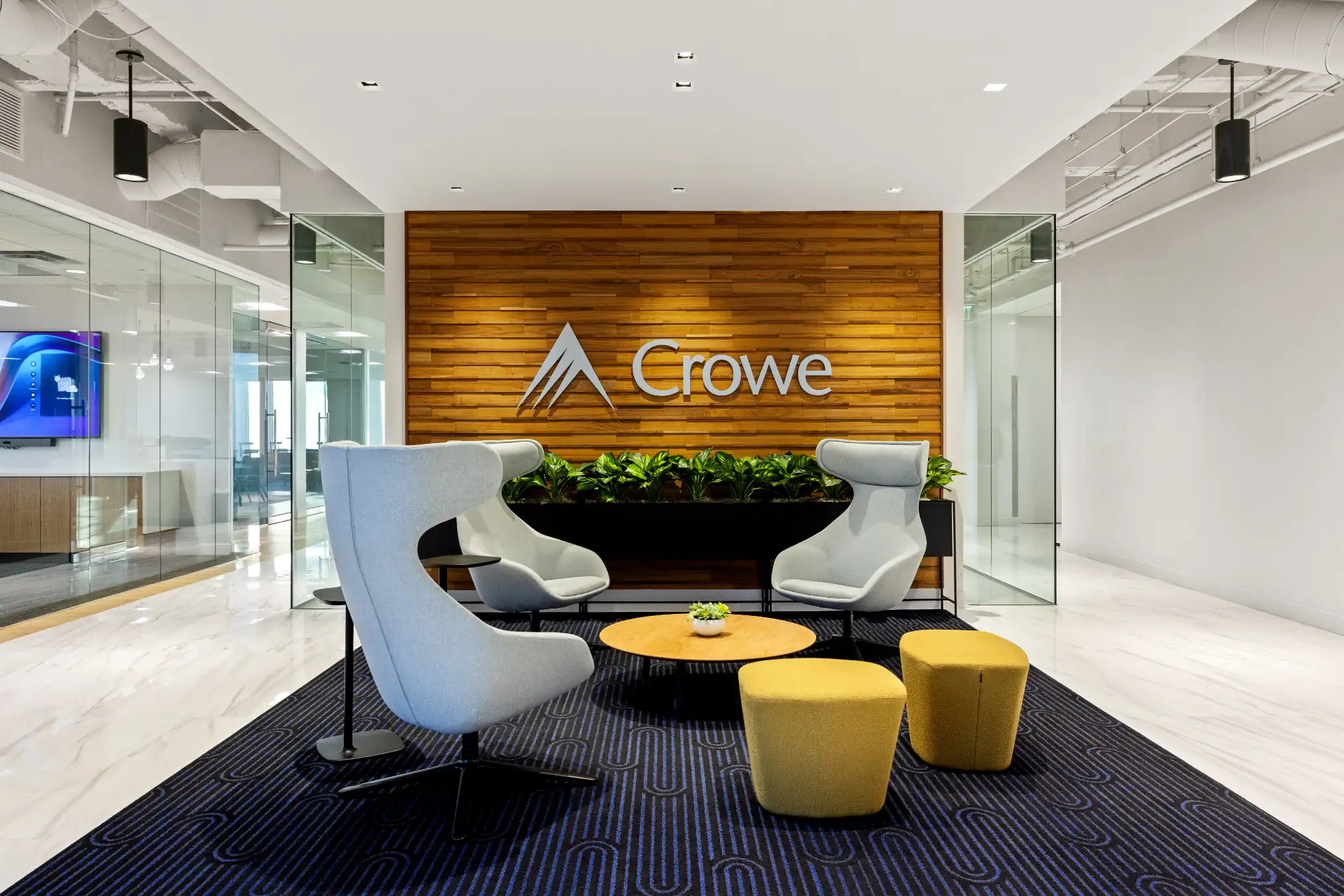 Modern office lobby with two gray lounge chairs, three yellow ottomans, a round wooden table, and a wooden wall featuring the Crowe logo, flanked by glass walls and green plants.