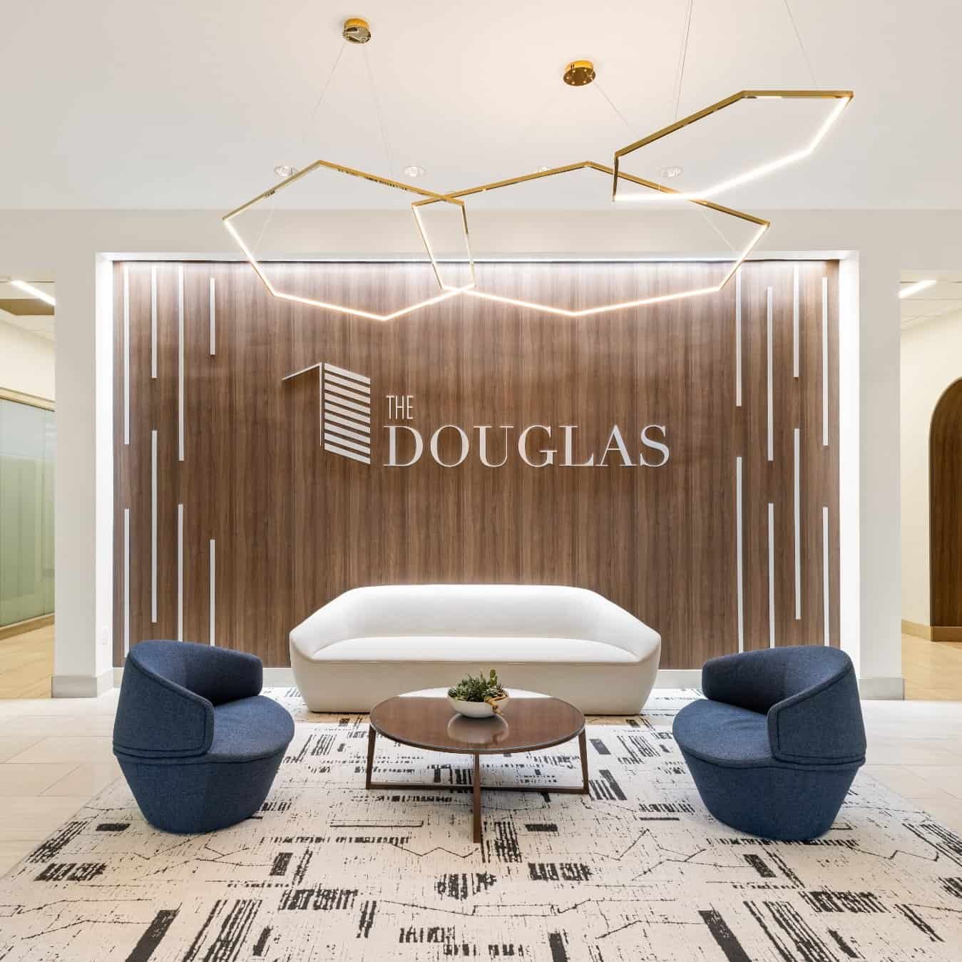 Modern lobby with a white sofa, three blue chairs, a round coffee table, and a geometric chandelier. A wood accent wall behind displays THE DOUGLAS in large white letters. Patterned rug on the floor.