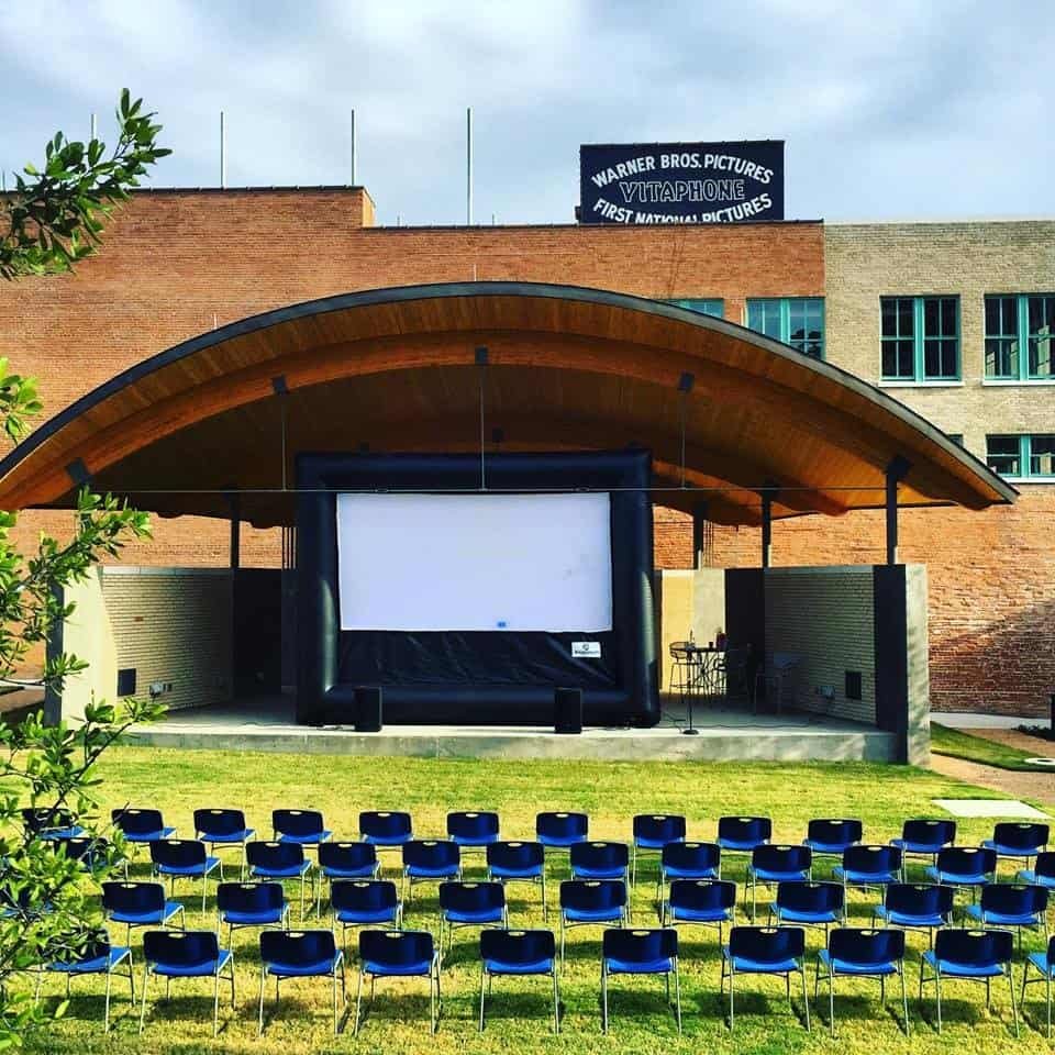 An outdoor stage with a large screen set up under a curved wooden roof, surrounded by brick buildings. Rows of blue chairs face the screen on a grassy lawn. A Warner Bros. Pictures sign is displayed above.