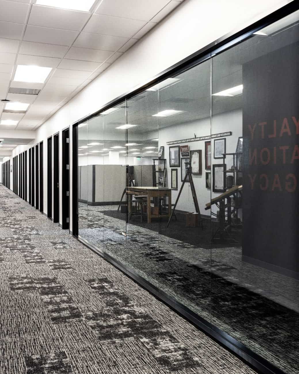 A modern office hallway with patterned carpet, closed doors on the left, and glass walls on the right revealing an office space with framed certificates, a desk, and equipment. The area is brightly lit with ceiling lights.