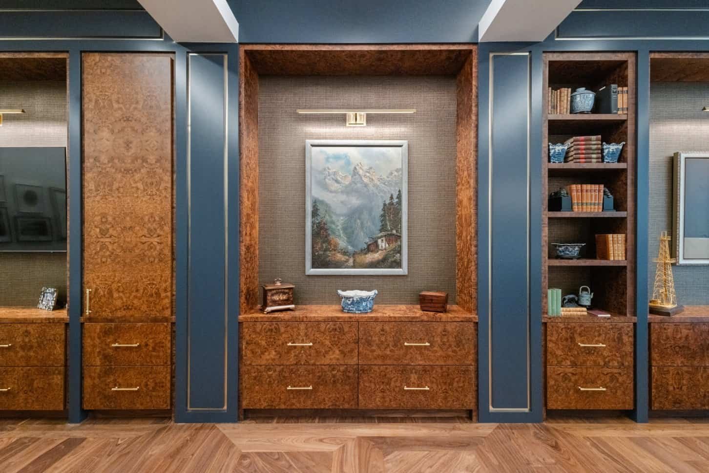 Built-in wood cabinets with gold handles surround a framed mountain landscape painting, flanked by bookshelves holding books and decorative items in a stylish, elegant room with herringbone wood floors.