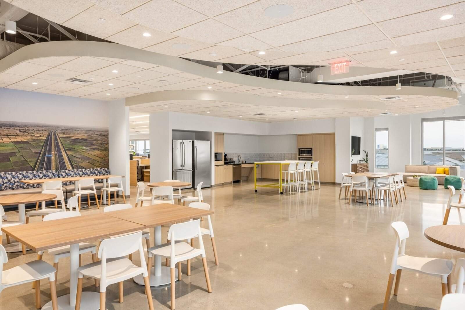 A modern, spacious office kitchen and dining area with wooden tables, white chairs, a kitchenette with appliances, a large wall photo, and lounge seating by large windows. The space is bright with natural and ceiling lights.
