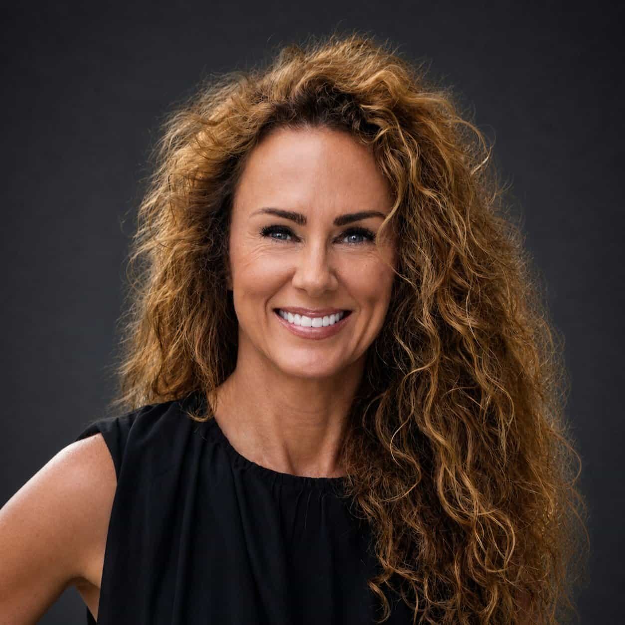 A woman with long, curly brown hair and a bright smile poses against a dark background, wearing a sleeveless black top.