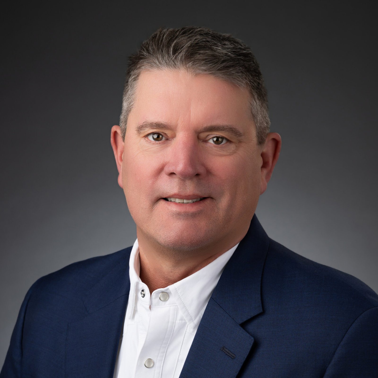 A middle-aged man with short gray hair, wearing a white button-up shirt and a dark blue blazer, poses against a dark gray background. He is facing the camera with a neutral expression.