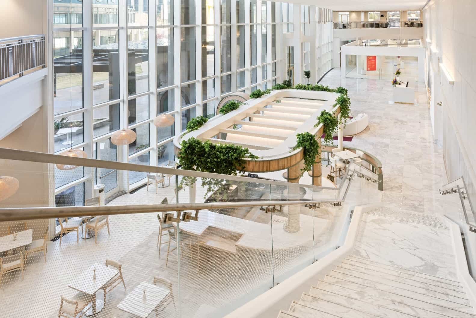 A bright, modern atrium with large floor-to-ceiling windows, white marble floors, a central seating area surrounded by greenery, and multiple tables and chairs. Wide stairs descend into the open, airy space.