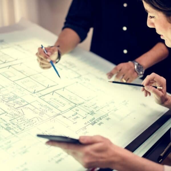 Two people study architectural blueprints on a large, illuminated table. One points with a pen while the other holds a tablet and pencil, both focused on the detailed floor plans in front of them.