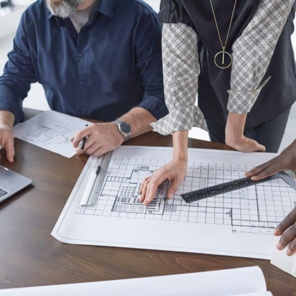 Three people collaborate at a desk, examining architectural blueprints. One person points at the plans while another uses a ruler. A laptop, documents, and drawing tools are also visible on the desk.