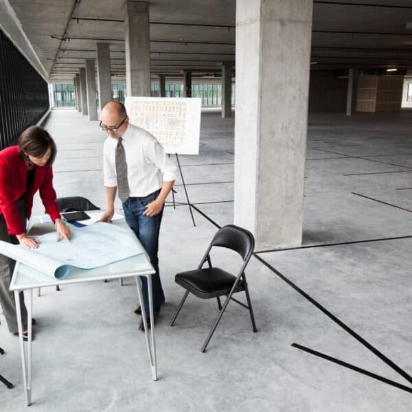 Two people stand at a table in a large, empty, unfinished office space, reviewing blueprints. Three chairs and a whiteboard with plans are nearby. The room has concrete floors, pillars, and large windows.